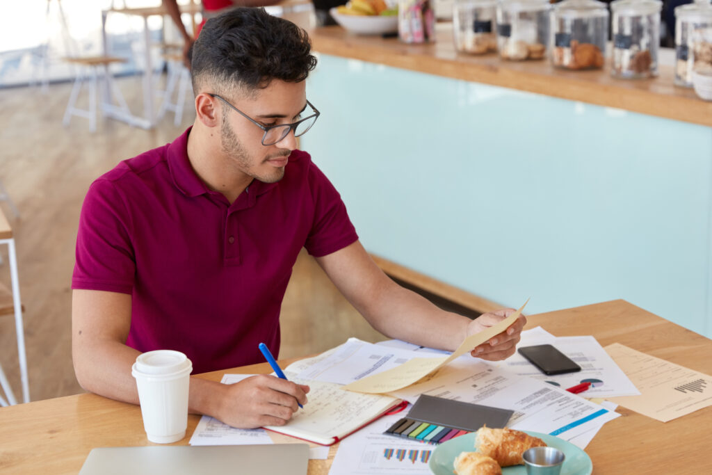 Busy male expert studies marketing issues, surrounded with documents, learns graphics and diagrams, uses stickies and notepad for noting information, spends lunch time in cafeteria or coffee shop